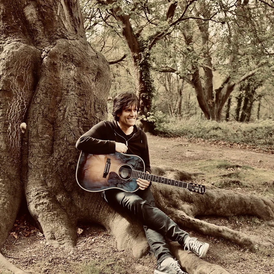 Lionel Fornetti sitting against a tree with an acoustic guitar, sepia tones