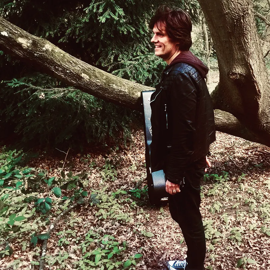 Lionel Fornetti in profile in the woods, guitar on his back