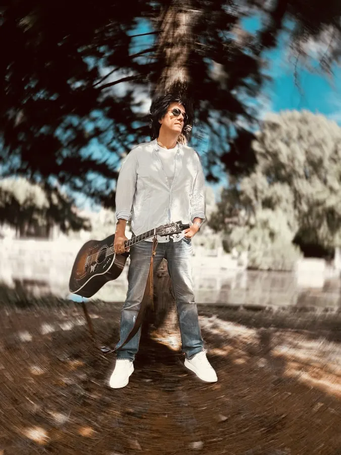 Lionel Fornetti standing with an acoustic guitar in a park, wide angle