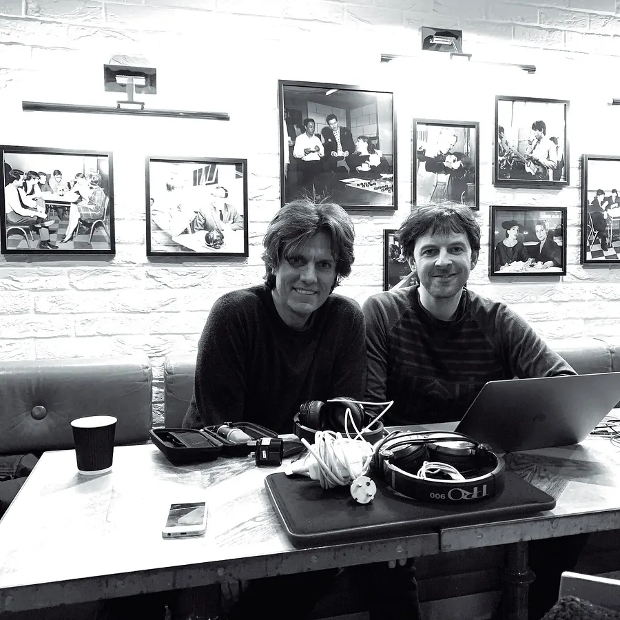 Two men at a table with a laptop, framed photos on the wall