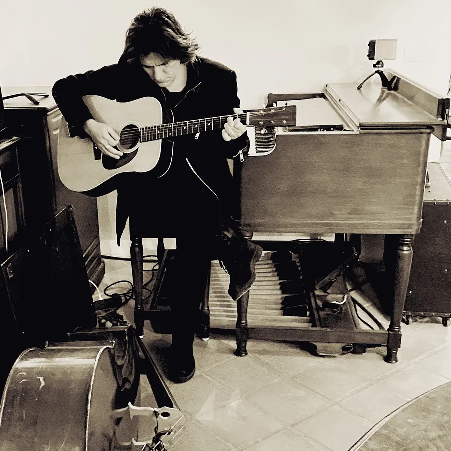 Lionel Fornetti playing acoustic guitar in a studio full of instruments, black and white